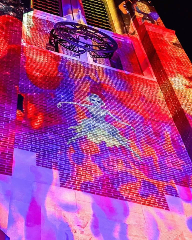 A close-up of the upper-section of Beaumont Tower displays bright red, violet, and yellow projections, centering on a glowing figure in motion beneath the black metal clock frame.