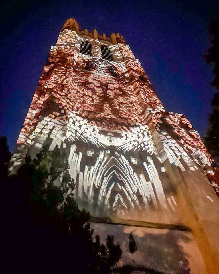 Beaumont Tower bathed in golden-white projections that form a cathedral-like arch pattern across the brick surface below the dark night sky.