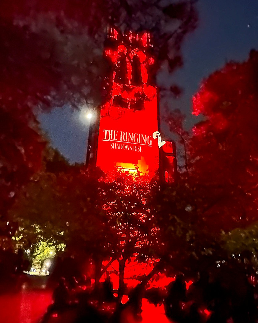A wide view of Beaumont Tower glowing entirely in red light, surrounded by trees. Projected text reads