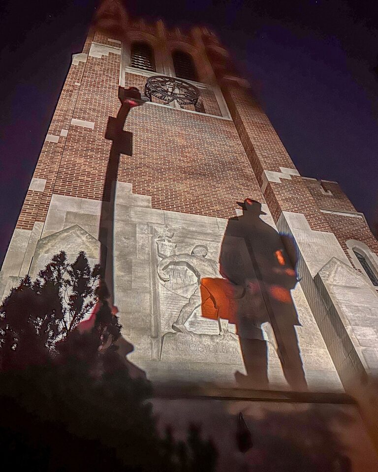 A night projection on Beaumont Tower showing the large shadow of a person in a hat across the lower facade, partially covering the tower's carved relief.