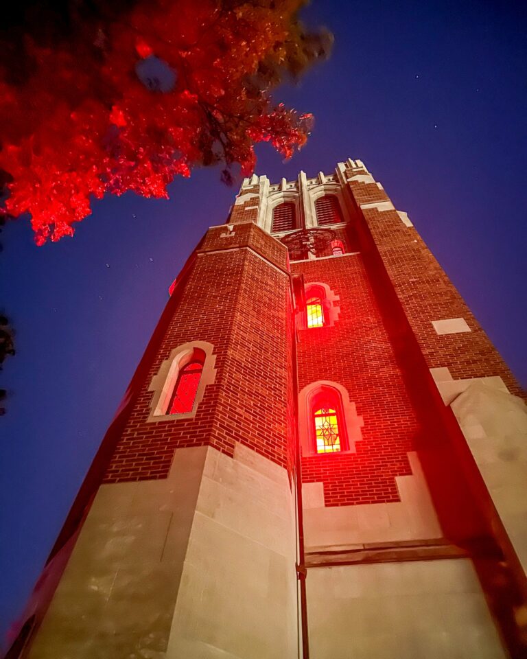Beaumont Tower viewed from below at dusk, its windows illuminated with red light that contrasts against the blue twilight sky and autumn leaves above.