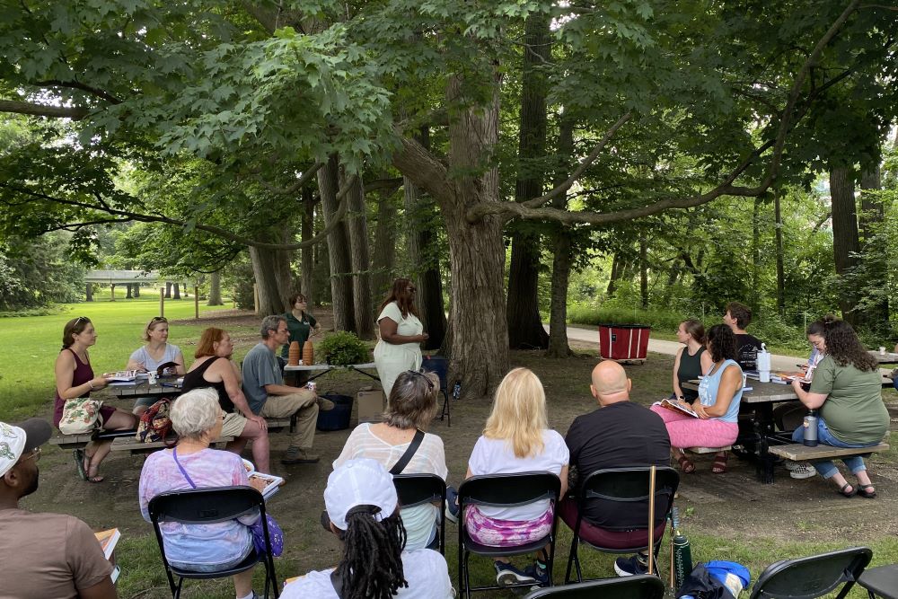 A group of more than 5 people sit at picnic tables and in chairs underneath trees in Beal Botanical Garden.