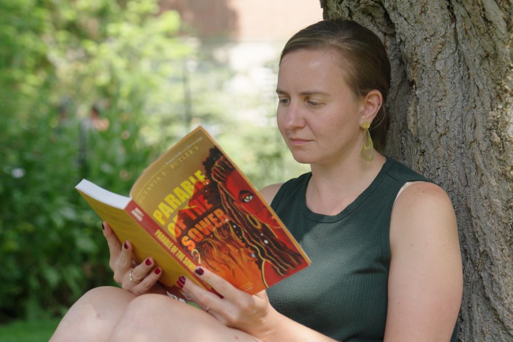 A person sits against the trunk of a tree and reads a book titled 
