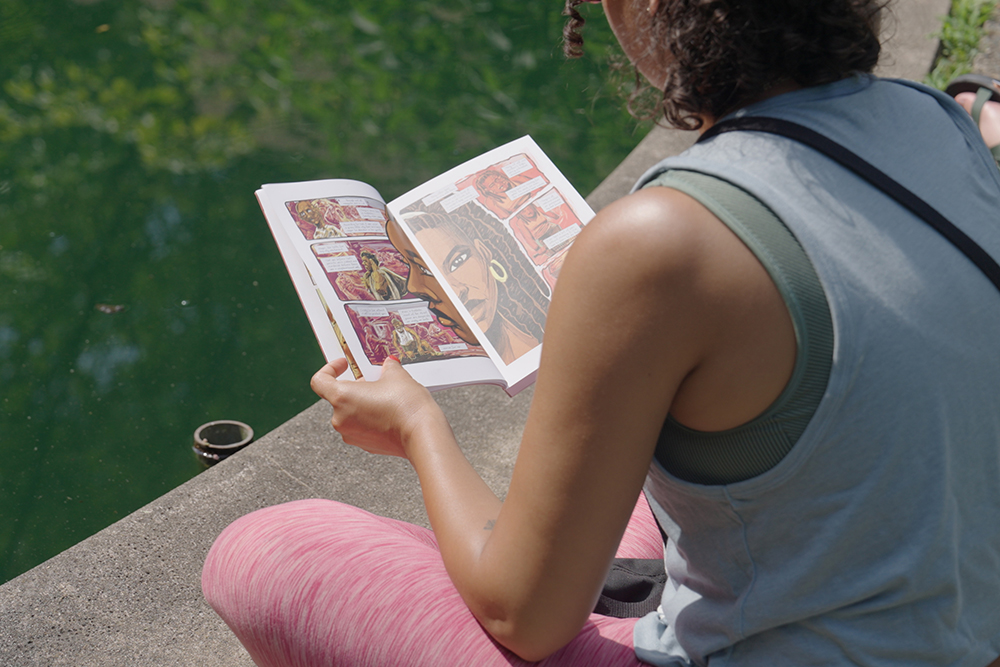 A person sitting next to a pond holds a graphic novel book and reads the pages.