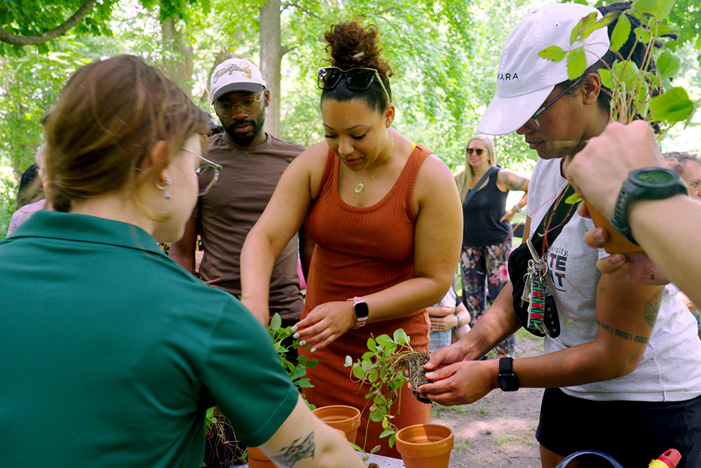 Two women, Kinitra Brooks and Angelica Bajos, stand next to each other smiling. In the background are green trees and foliage.