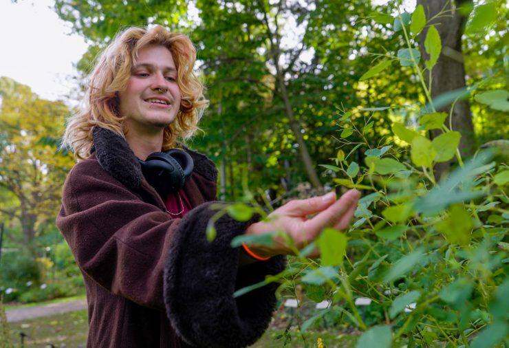 A person stands outside surrounded by greenery. They are wearing a maroon jacket and reaching out their hand to touch a plant.