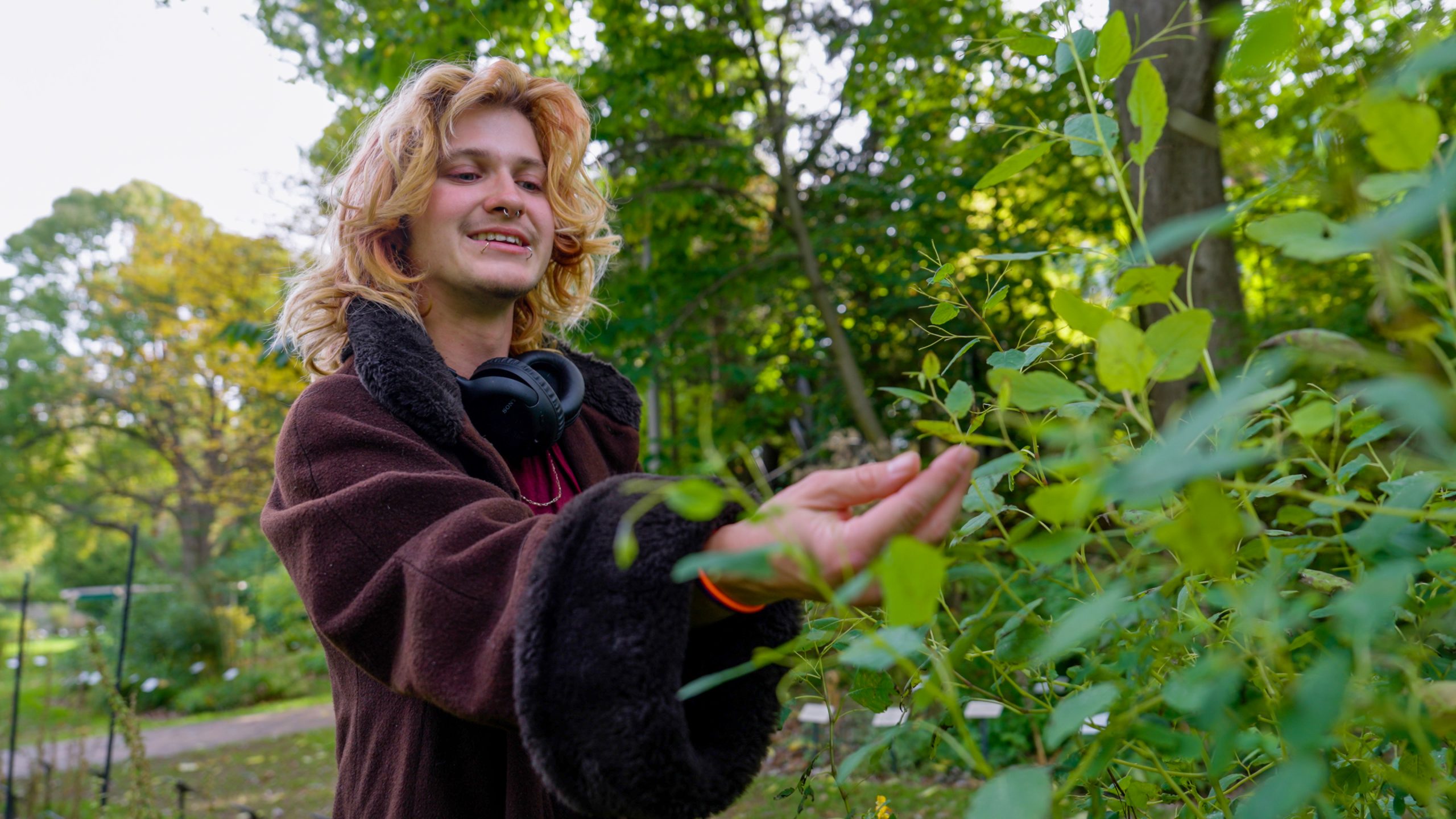 A person stands outside surrounded by greenery. They are wearing a maroon jacket and reaching out their hand to touch a plant.