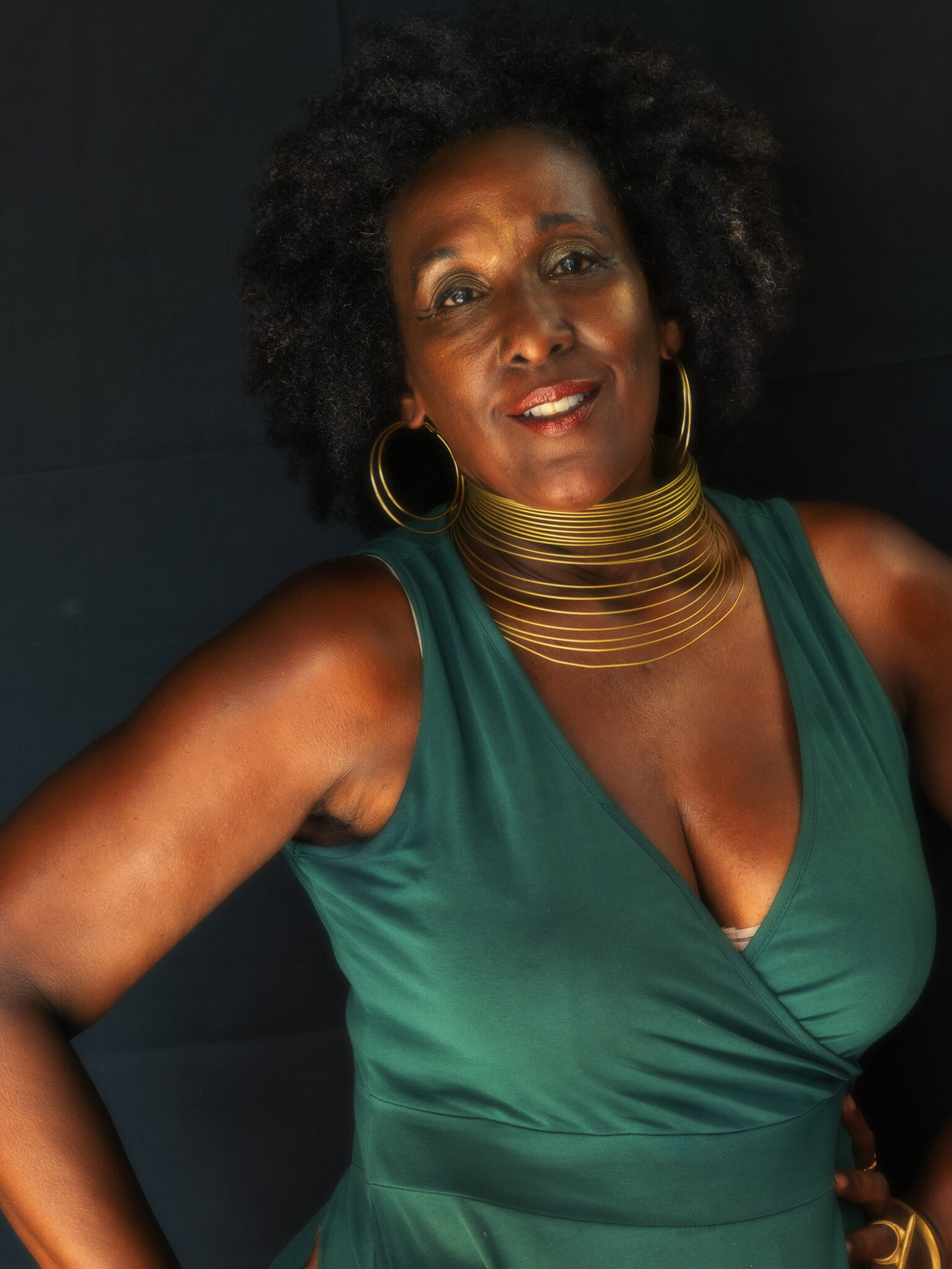 Woman in green tank top and gold choker necklaces and hoop earings looks at camera