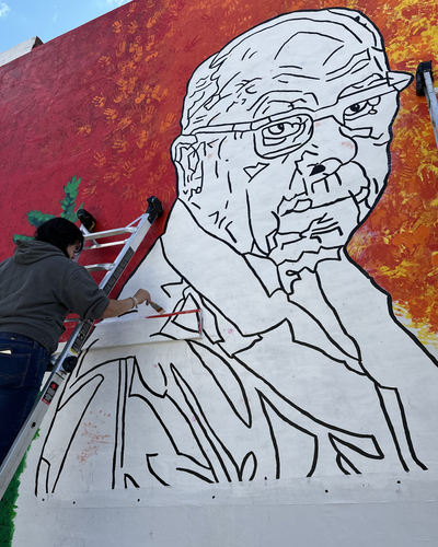 A person stands on a ladder holding a paintbrush. They are painting part a mural depicting a man wearing a suit and glasses.