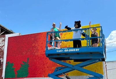 Three people including one in a bear mascot costume, stand on a blue lift waving in front of a red, orange, and yellow mural in progress on the side of a brick building under a clear blue sky.