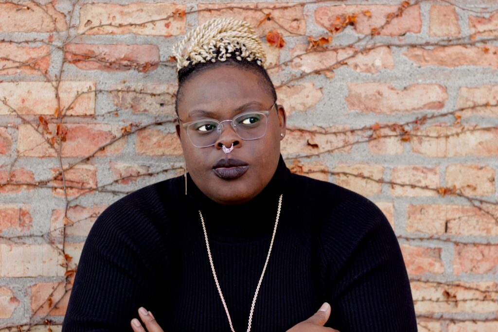 Woman with white twists piled into a top bun, wearing a black turtleneck and clear glasses with septum piercing