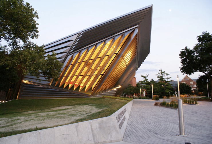 A metallic building with trees surrounding it and sidewalk leading up to the entrance. It is evening and yellow light is pouring out the windows of the front of the building.