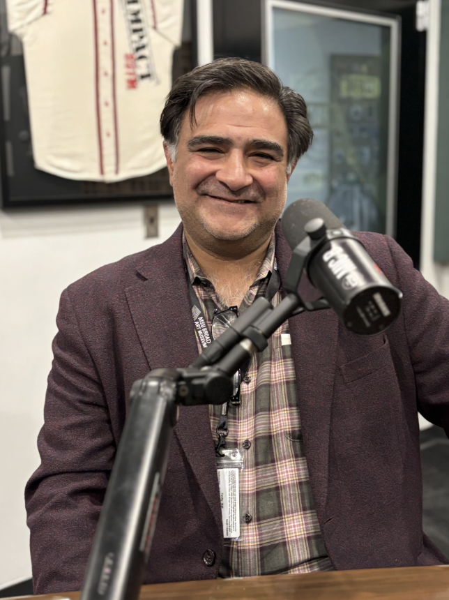A man sits smiling at a table with a microphone in front of him. He is wearing a maroon blazer and a plaid button up shirt.