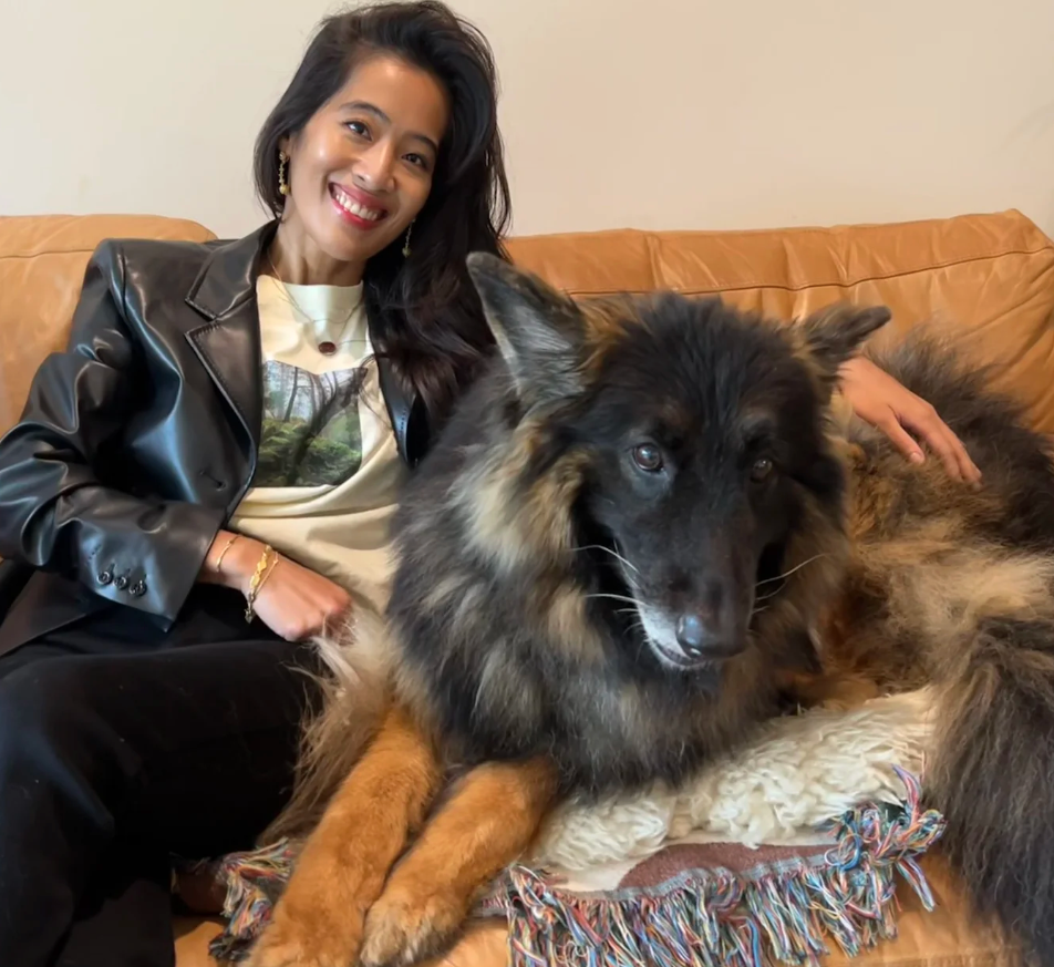 Woman with black hair and leather jacket sits on tan sofa with large german shepherd