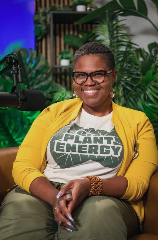 Woman with short hair, glasses, and yellow cardigan sits in front of a studio microphone