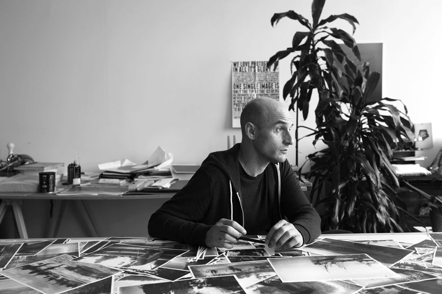 Black and white photo of man wearing black hoodie at desk with photos in front of him and a plant behind