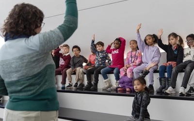 A teacher stands in front of a group of young students siting on tiered benches, engaging them in a lesson.