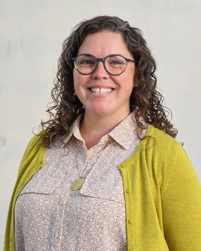 A woman with curly brown hair and glasses smiles while standing against a light grey background.