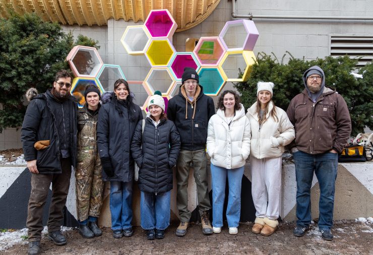 Eight people stand in line in front of the completed outdoor sculpture, which features interconnected pastel and translucent hexagon modules. They are dressed in winter coats, hats, and gloves, standing near shrubs and a building wall.