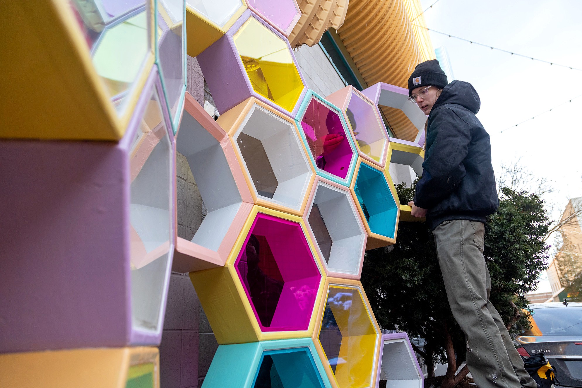 A person in a winter jacket and hat works beside the installed sculpture, reaching into one of the colorful hexagonal modules.The structure's translucent panels reflect the surrounding area as it rises vertically along the wall and into a cluster of evergreen branches.