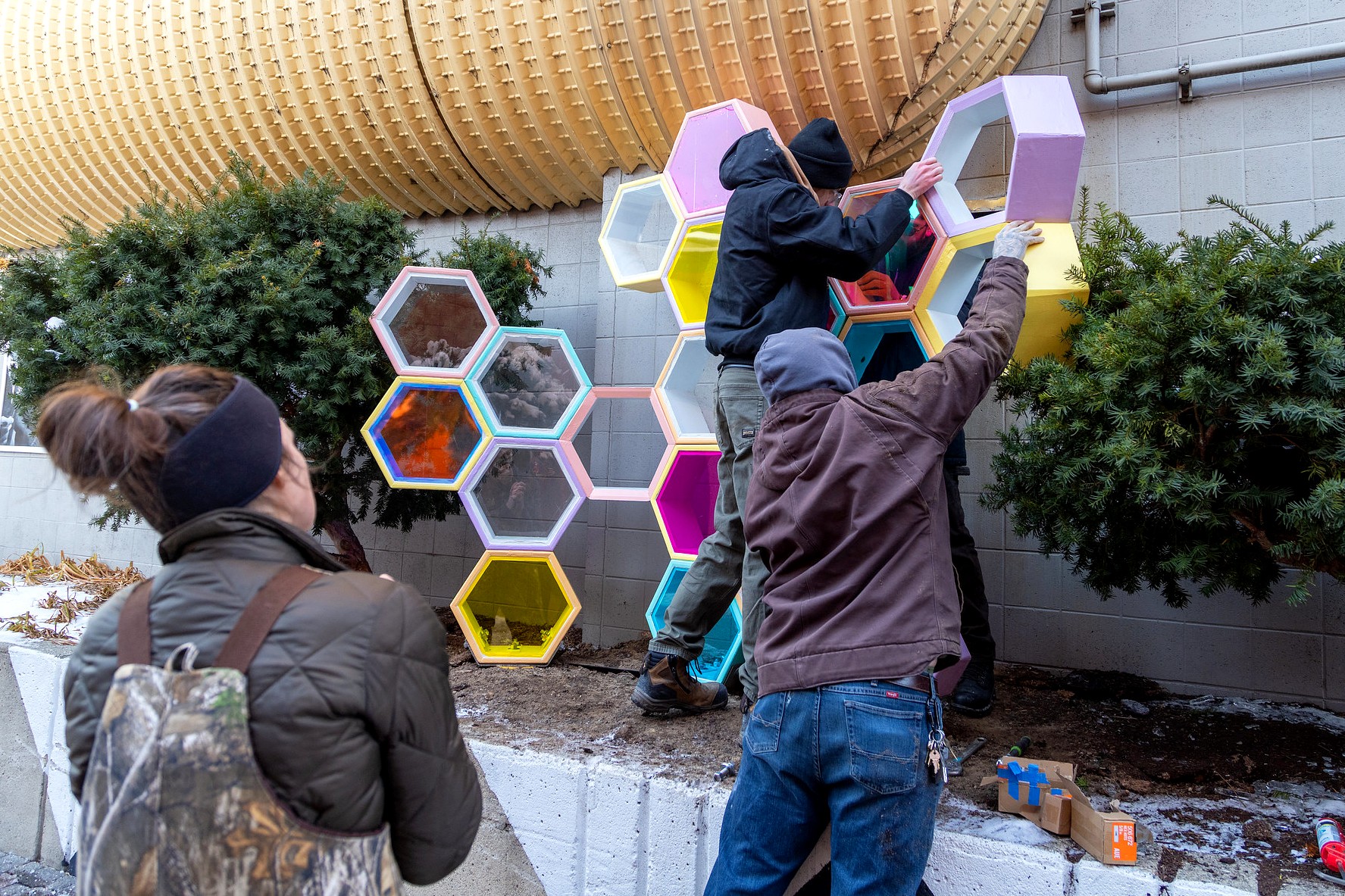 Three people work together to lift and position a section of pastel-colored hexagonal modules as part of the sculpture installation. One person reaches upward to steady the structure while another supports it from below. A fourth person observes from nearby.