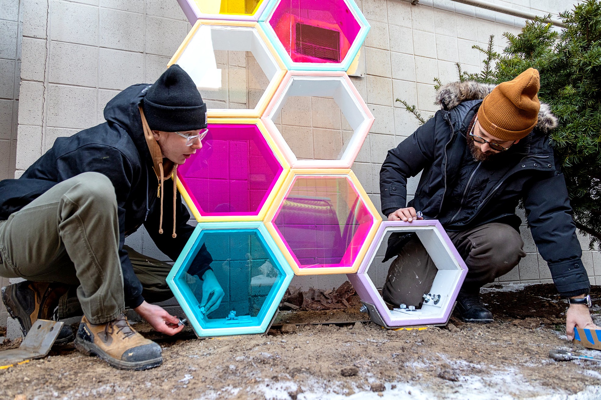 Two people crouch at the base of a sculpture, attaching bolts and hardware to secure the lowest hexagonal modules.