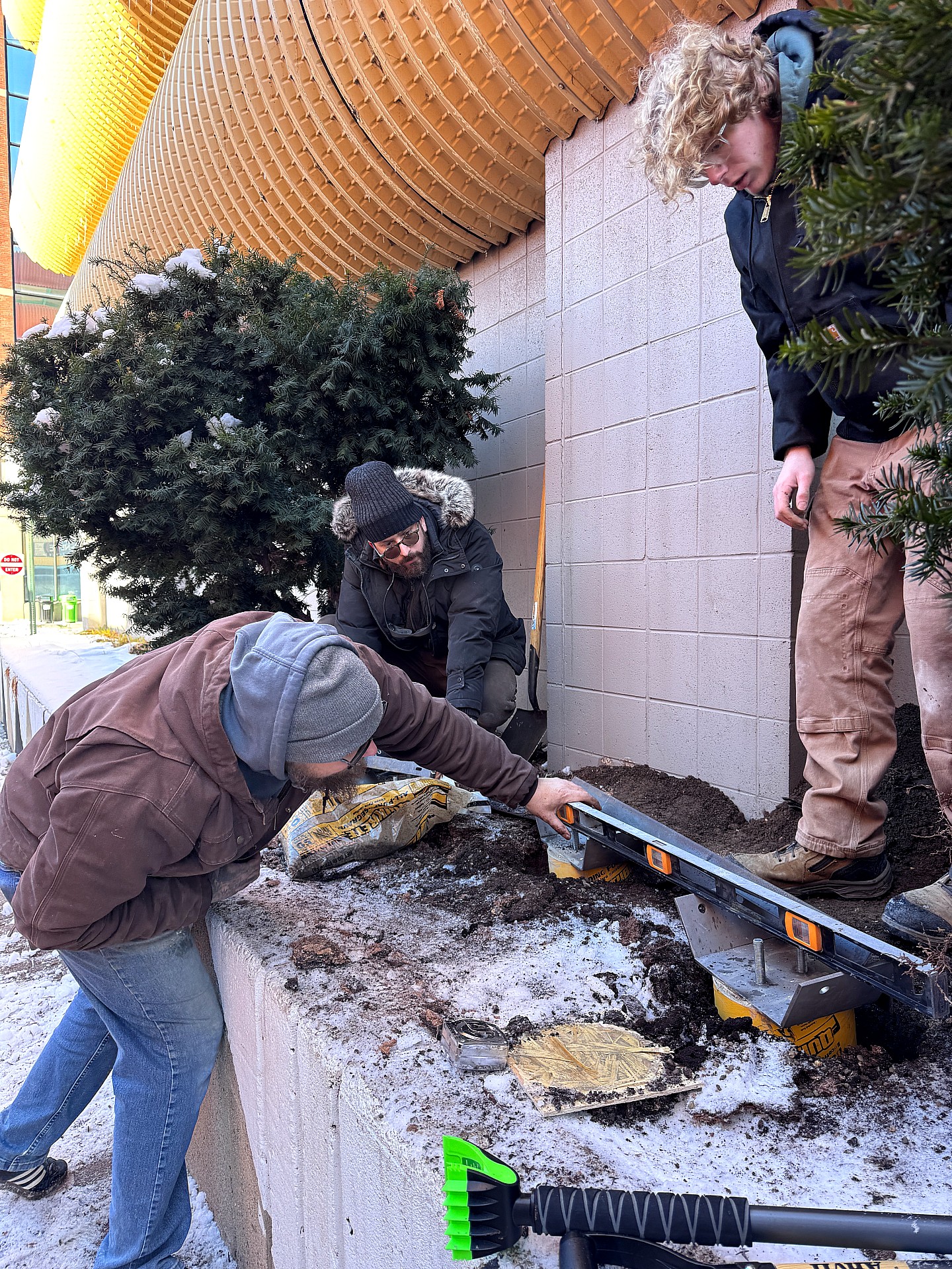 Three people prepare the installation site by leveling foundation supports on a snowy concrete ledge beside a building. They work with a long level, and tools while standing near evergreen bushes and the building's yellow overhang.