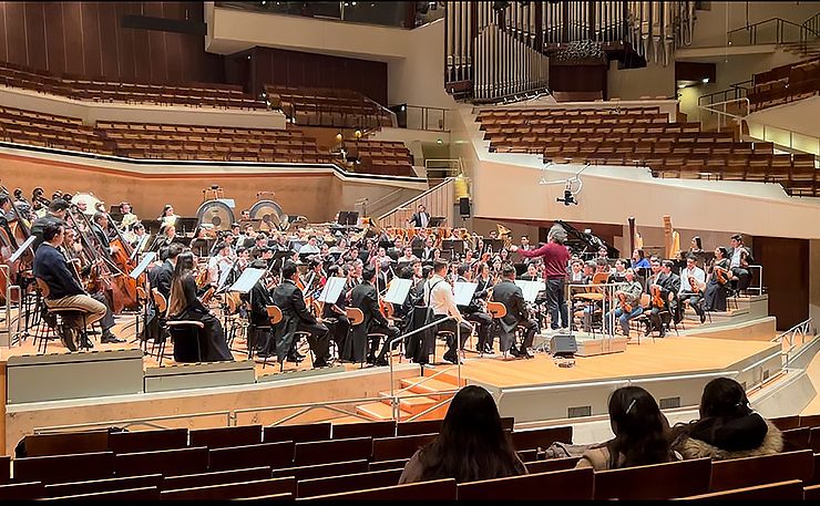 A full orchestra surrounds a conductors wearing a red sweater. Three women are seated in front of the stage looking ahead to the musicians.