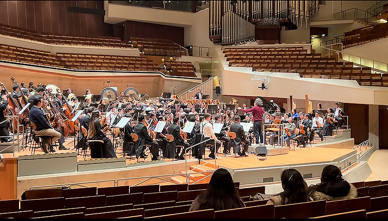 A full orchestra surrounds a conductors wearing a red sweater. Three women are seated in front of the stage looking ahead to the musicians.