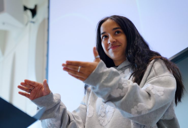 A woman with dark hair stands in front of a music stand. Her hands are raised in front of her as she gestures in a conducting pattern.