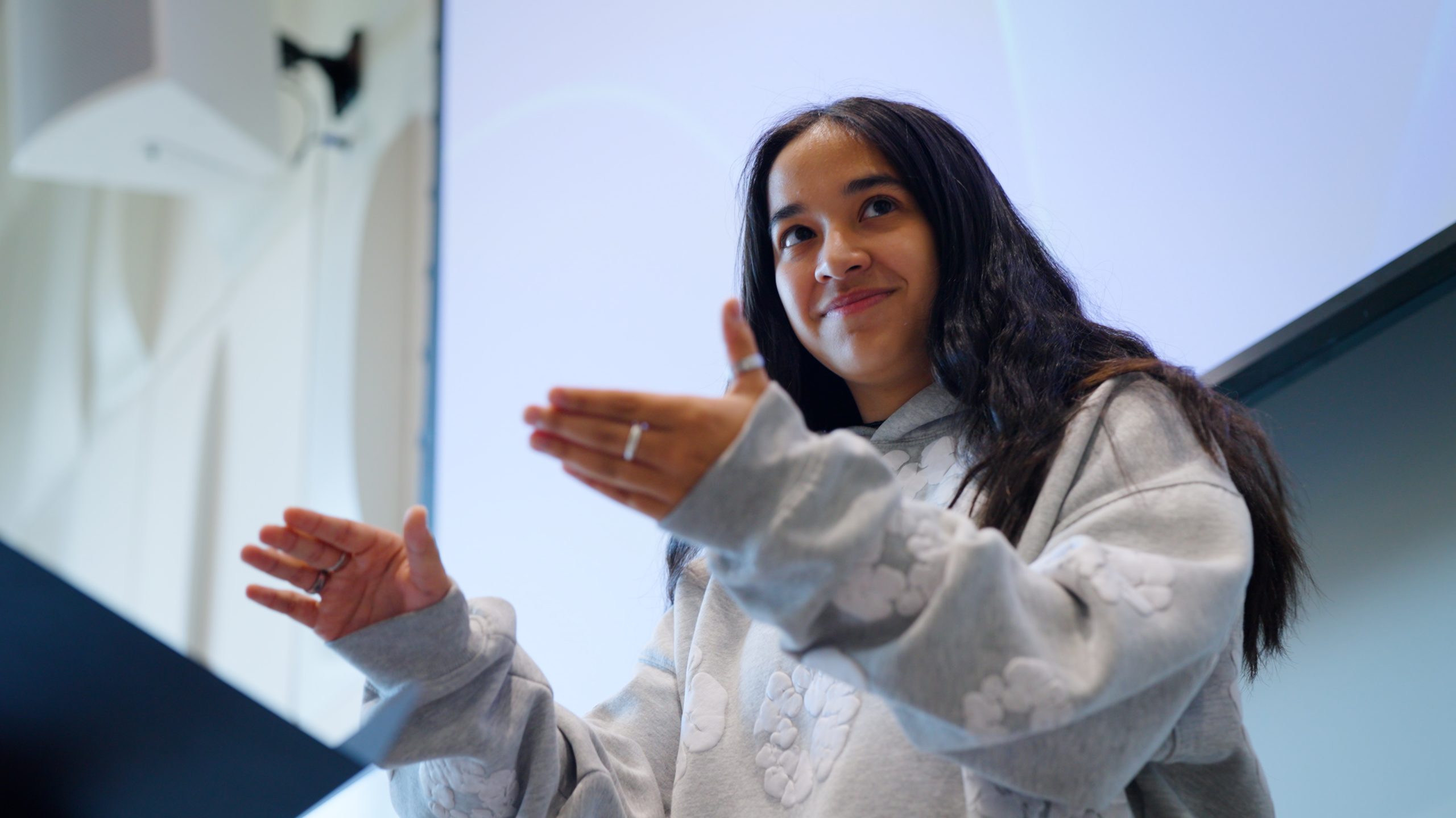 A woman with dark hair stands in front of a music stand. Her hands are raised in front of her as she gestures in a conducting pattern.