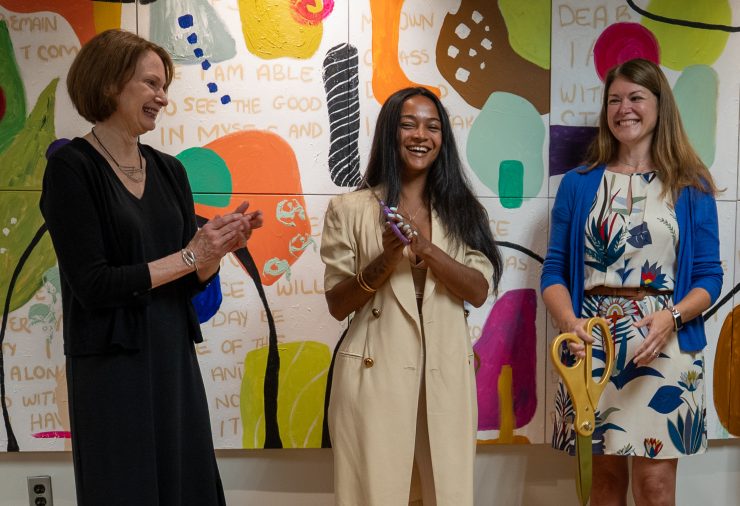 Three woman stand in front of a colorful mural that is painted with various shapes and words. The woman on the left wears a long black dress and is smiling and clapping. The woman in the center wears a long tan coat with gold buttons, she is smiling with her hands clasped in front of her holding a pair of purple scissors. The woman on the right wears a tan dress with blue plants, and a blue cardigan. She is smiling and holding a comically large pair of gold scissors.