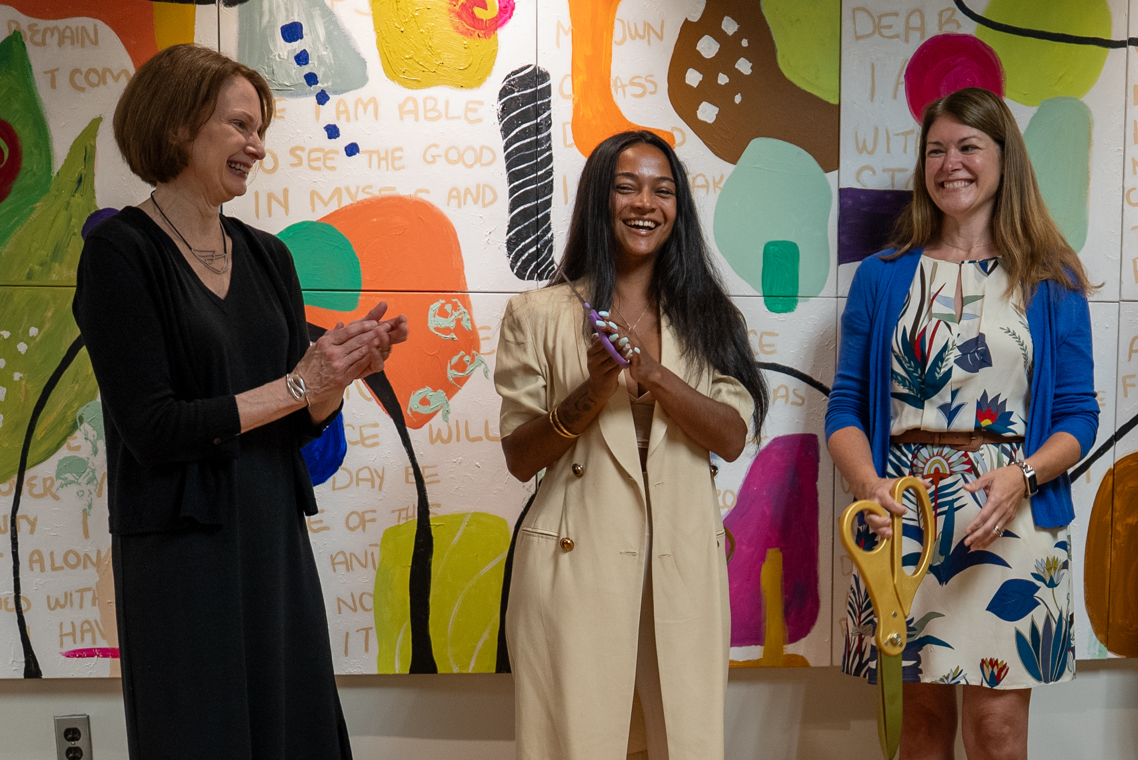 Three woman stand in front of a colorful mural that is painted with various shapes and words. The woman on the left wears a long black dress and is smiling and clapping. The woman in the center wears a long tan coat with gold buttons, she is smiling with her hands clasped in front of her holding a pair of purple scissors. The woman on the right wears a tan dress with blue plants, and a blue cardigan. She is smiling and holding a comically large pair of gold scissors.
