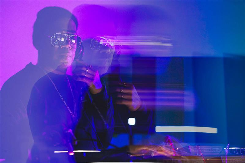 Blurred purple toned image of man with glasses and beanie DJing