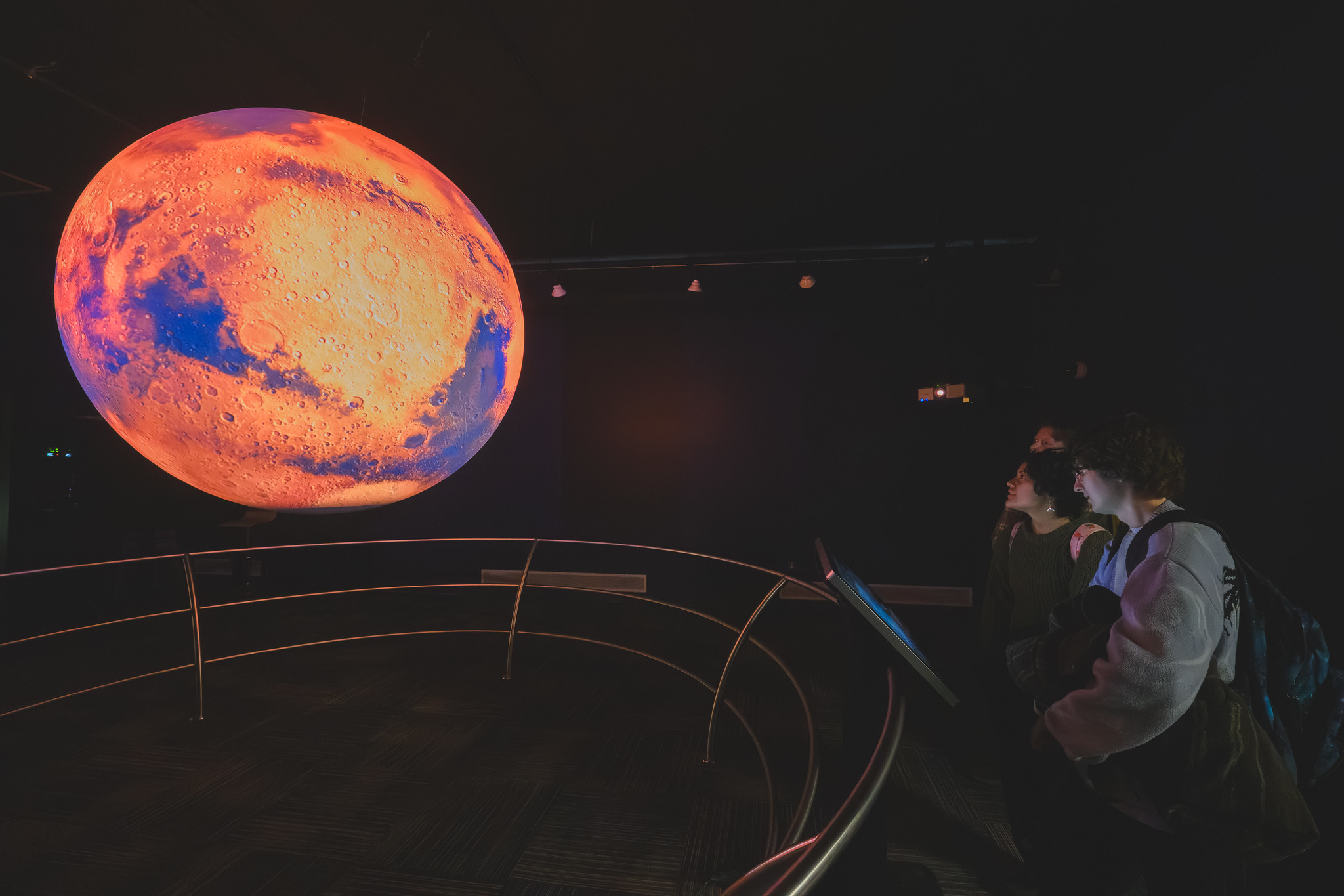 Three students in a dark room look up at a glowing blue and orange sphere depicting the earth's temperature