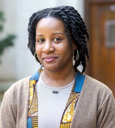 Portrait of a woman with shoulder-length twisted braids wearing a light-colored top, patterned trim, and a brown cardigan, standing indoors with a softly blurry background.