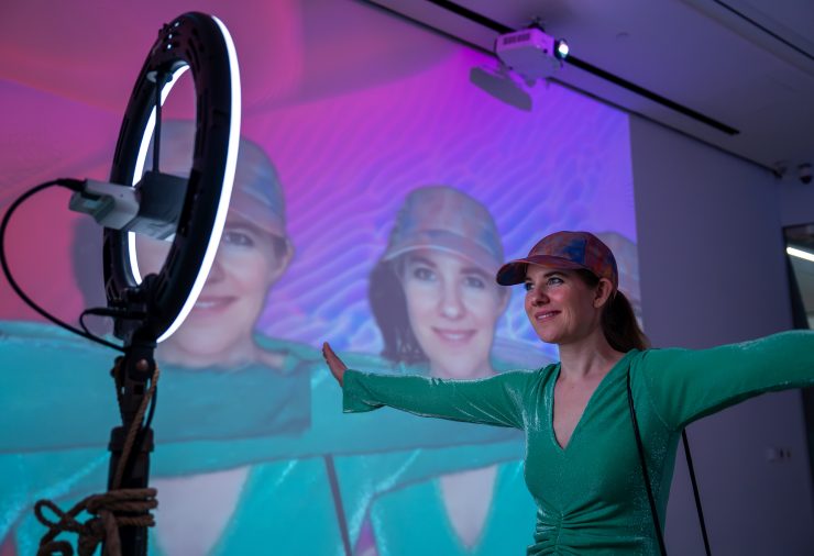 A person in a long dress stands in a dim gallery space, interacting with a digital art installation. A ring light on a stand faces them, while large projections of the person's face cover the walls.