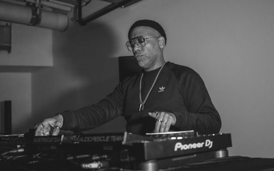 A DJ performs in a dimly lit room, standing behind a set of Pioneer turntables and audio equipment. They wear a dark long-sleeve shirt with a small logo and a chain necklace, with one hand adjusting controls on the mixer. The scene is captured in black and white, highlighting the industrial ceiling pipes above and the focused atmosphere of the performance.
