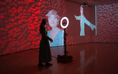 A person in a long dress stands in a dim gallery space, interacting with a digital art installation. A ring light on a stand faces them, while large projections cover the surrounding walls–one showing a textured red background and another displaying a life-sized figure mimicking their pose. The polished wooden floor reflects the glowing colors of the immersive environment.