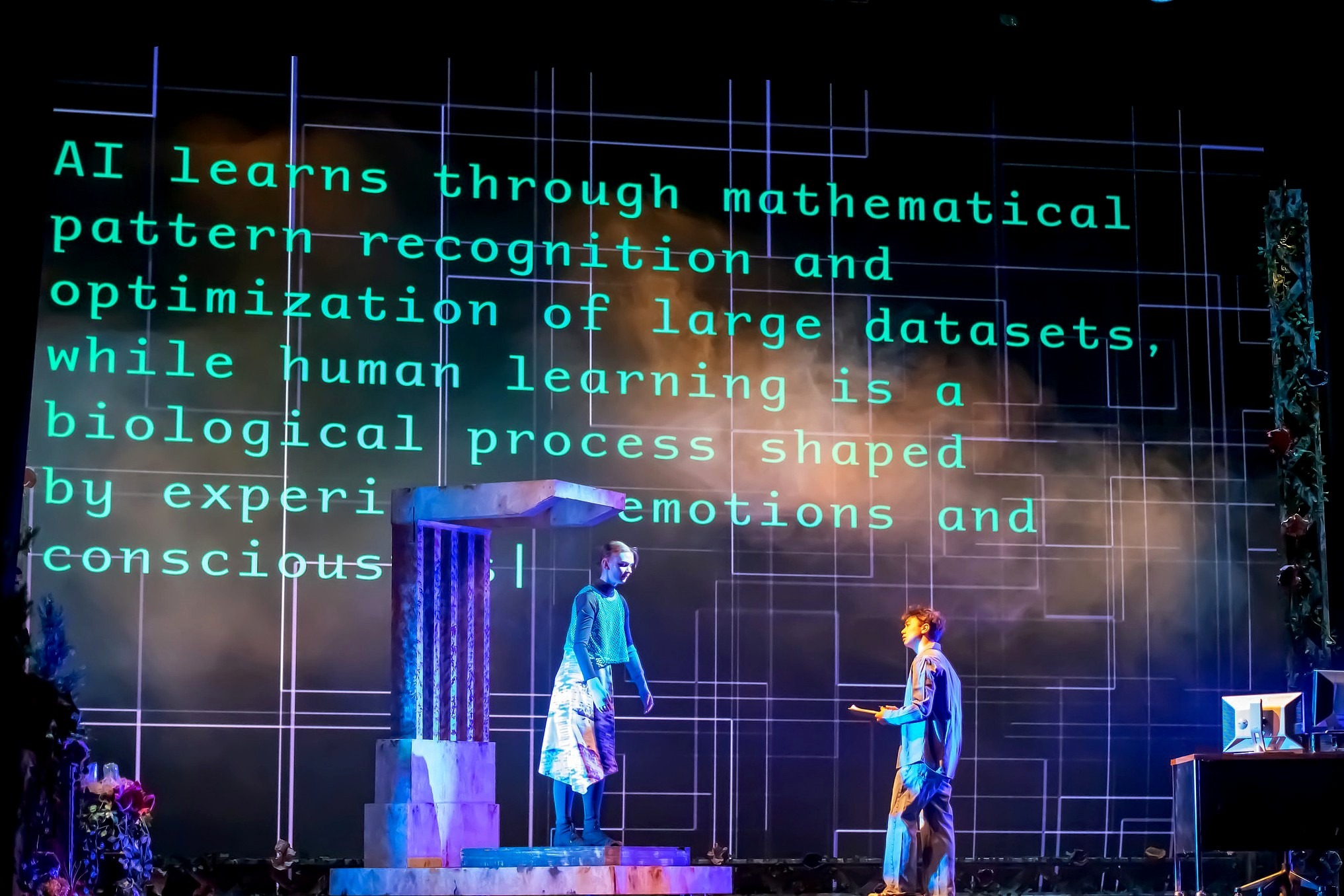 Two student performers stand on stage beneath a large projection reading. 
