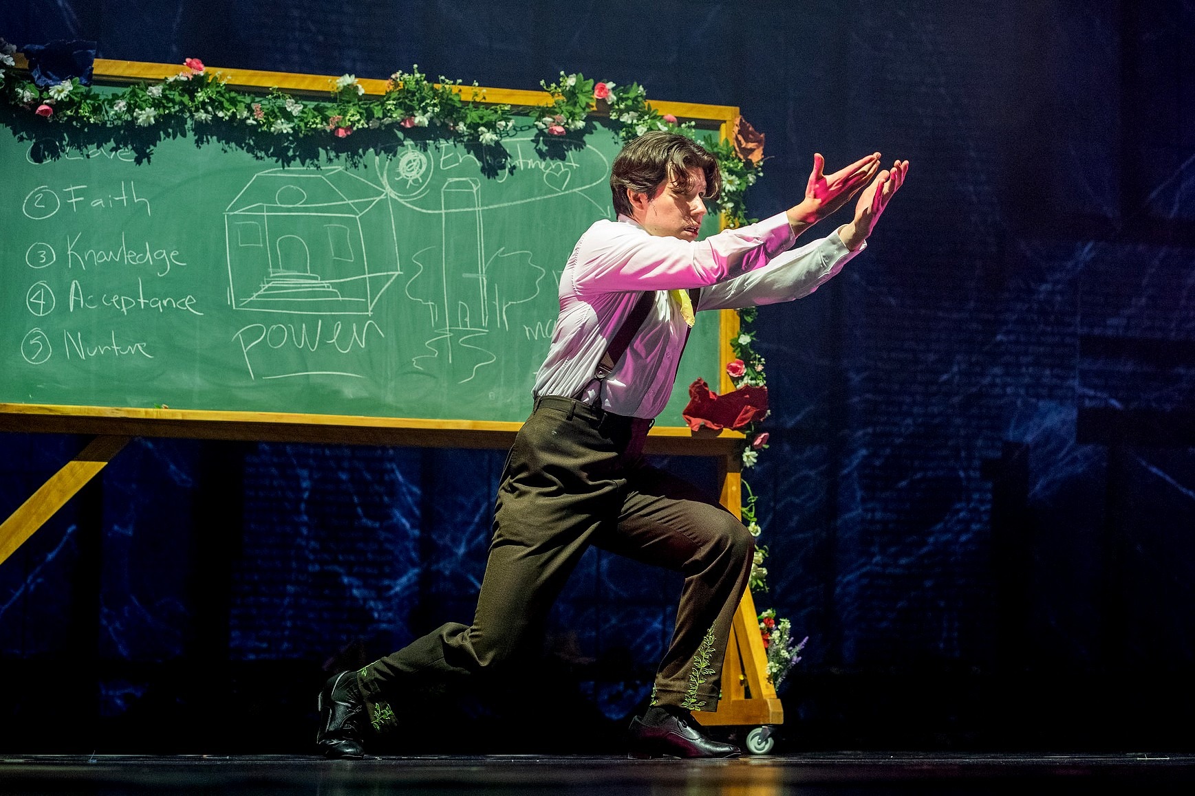 A dancer lunges forward in a dramatic pose before a chalkboard decorated with garland. The board displays hand-drawn notes and images, including a building sketch and the words 