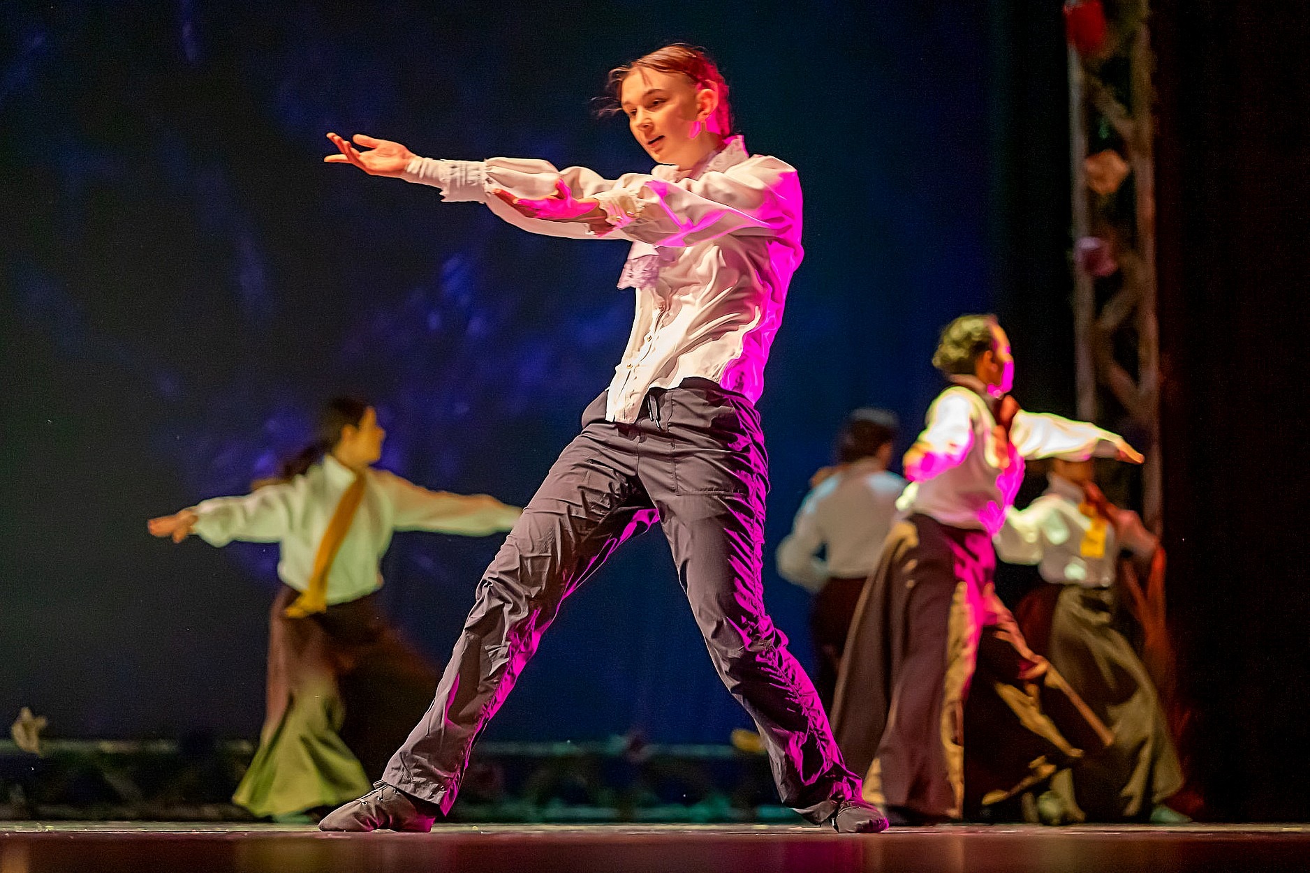 A student dancer performs a wide, grounded stance with arms extended under pink and blue stage lighting. Additional performers move in the background.