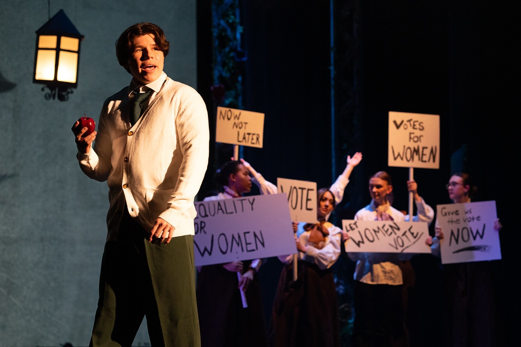 A performer stands beneath a stage lantern holding a red apple, while a group of student performers behind hold protest signs reading 