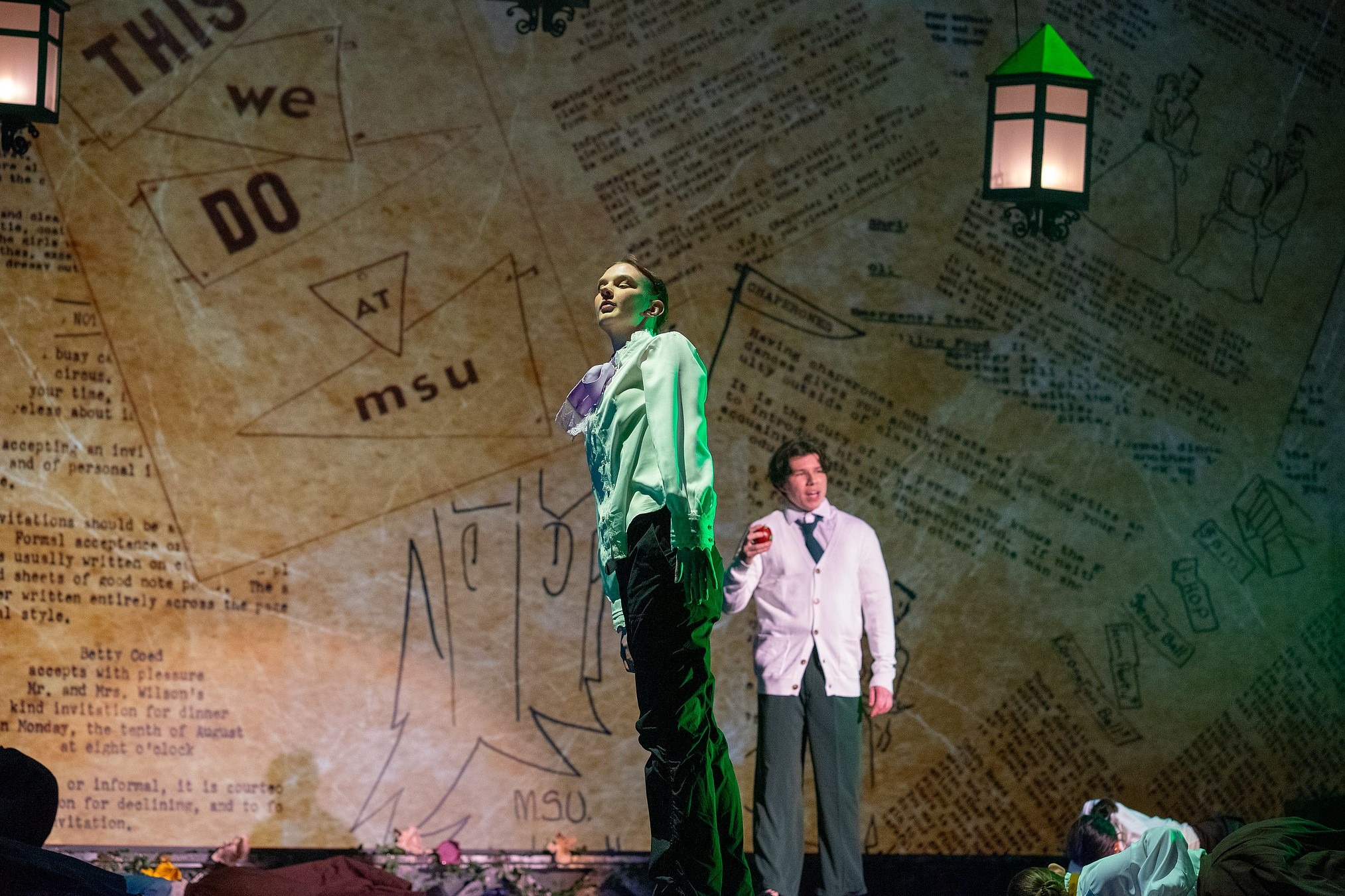A performer stands under green-tinted stage lighting in front of a backdrop designed like aged paper filled with text and diagrams, including the words 