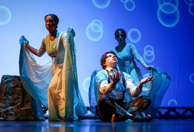 A performer sits cross-legged at center stage with palms raised upward, illuminated by cool blue lighting and circular projections. Two dancers stand behind holding flowing blue fabric.