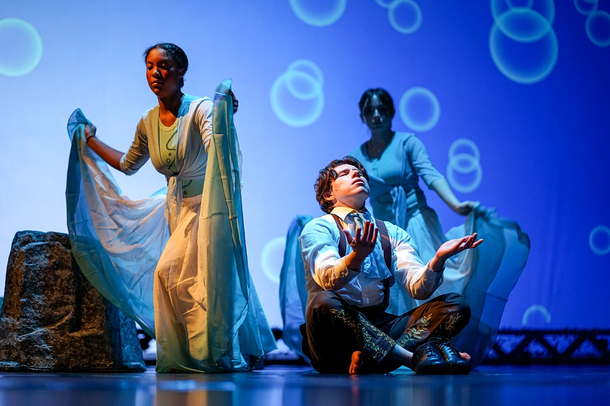 A performer sits cross-legged at center stage with palms raised upward, illuminated by cool blue lighting and circular projections. Two dancers stand behind holding flowing blue fabric.