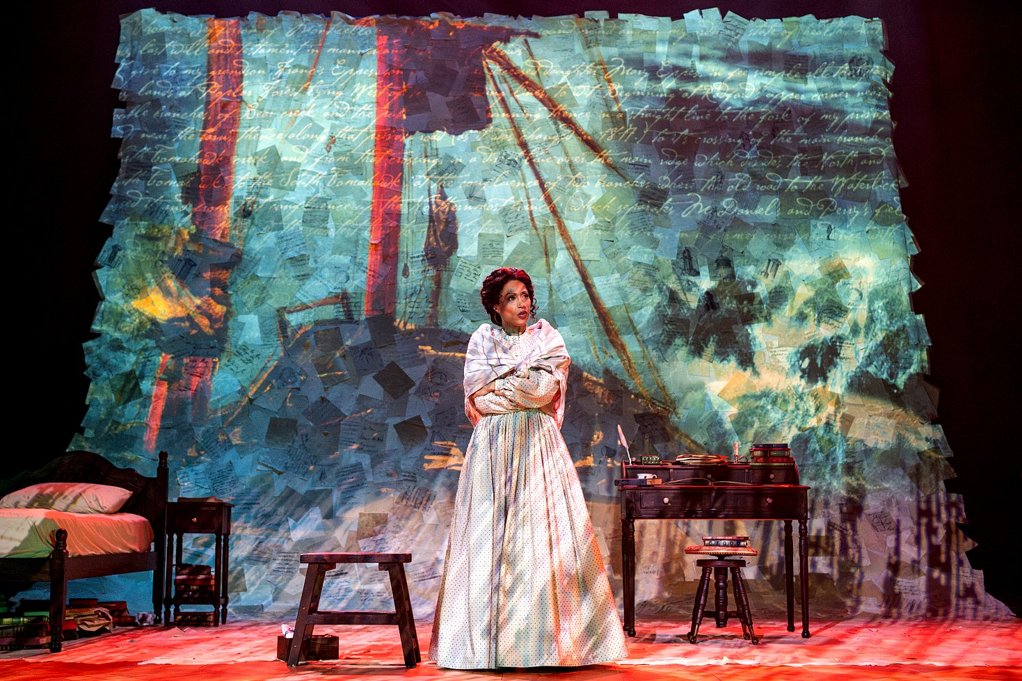 An actor stands center-stage in a cream-colored gown and a light-colored shawl wrapped around her shoulders. She has her arms crossed. The background features a large wall-sized projection of a 19th-century manuscript with red and yellow vertical highlights over a white script. A wooden stool and roll-top desk flank the actor on a stage floor lit with warmed light.