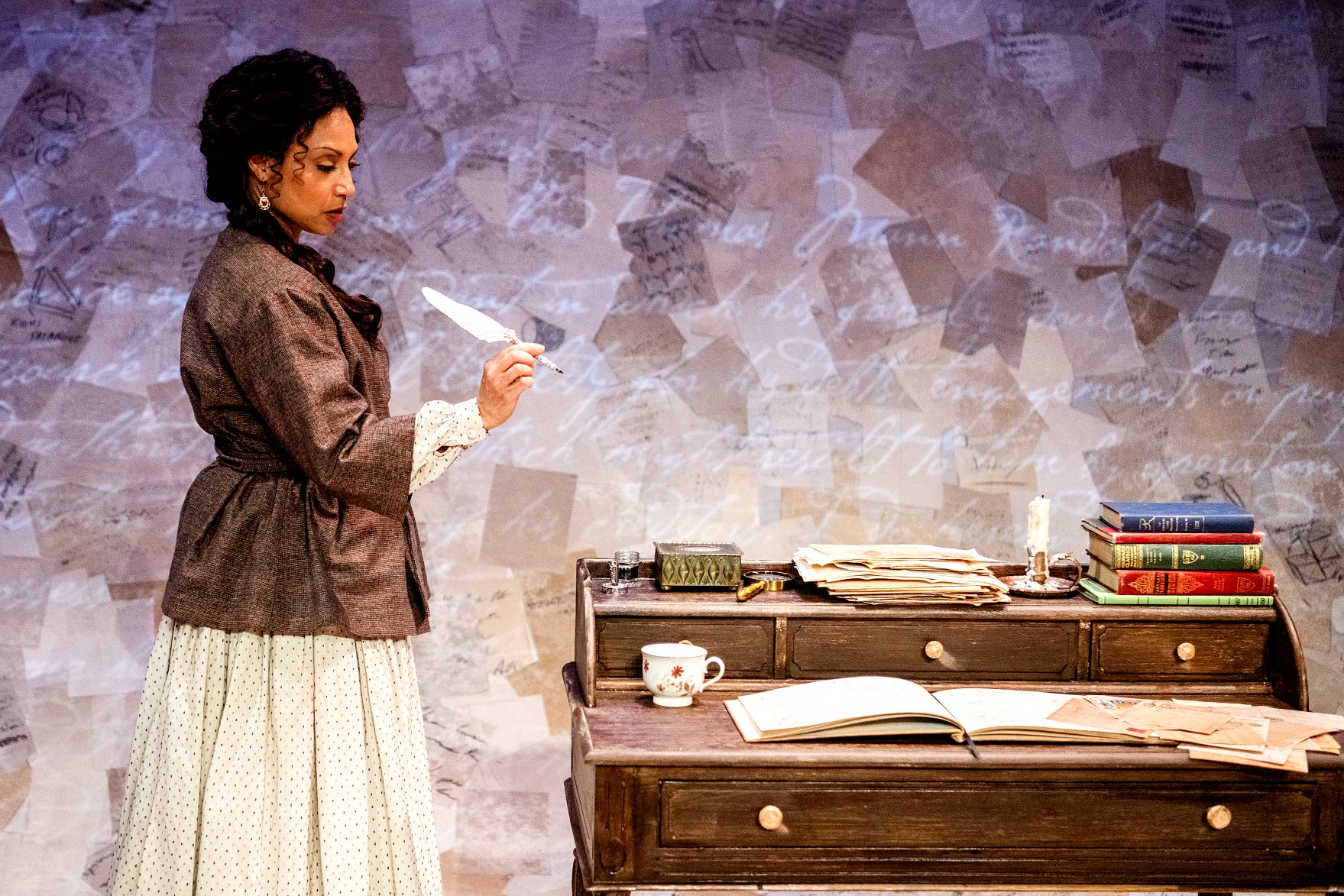 A woman in a mid-19th-century cream-colored skirt and a structured brown jacket stands at a wooden roll-top desk. She holds a white quill pen over a collection of parchment papers. The background features a large, textured wall projected with overlapping historical documents.