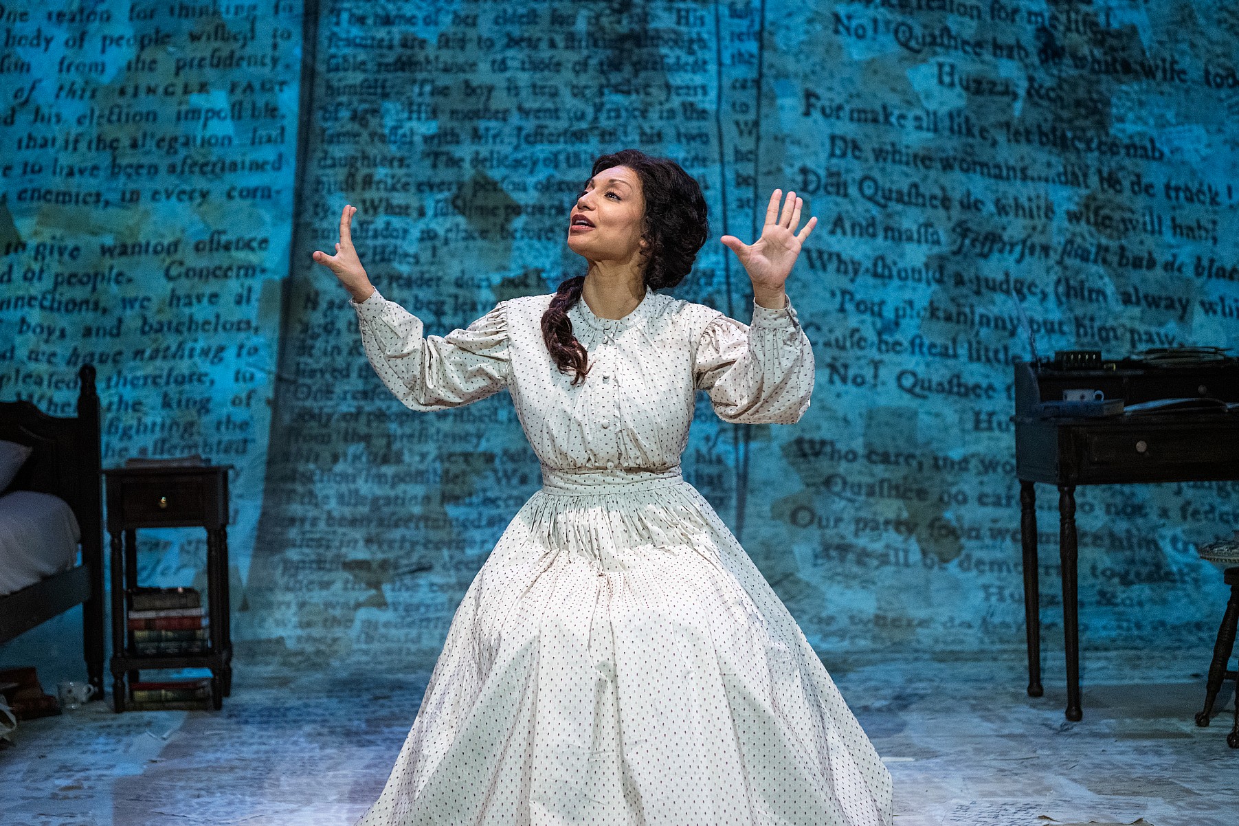 An actor is seated center stage, wearing a long-sleeved, high-collared cream colored period dress. Her hands are raised in an expressive, open gesture as she looks upward. The backdrop is a floor-to-ceiling projection of a historical newspaper text in a deep indigo blue. A small wooden nightstand with stacked books sits to the left and a desk with various papers sits to the right.