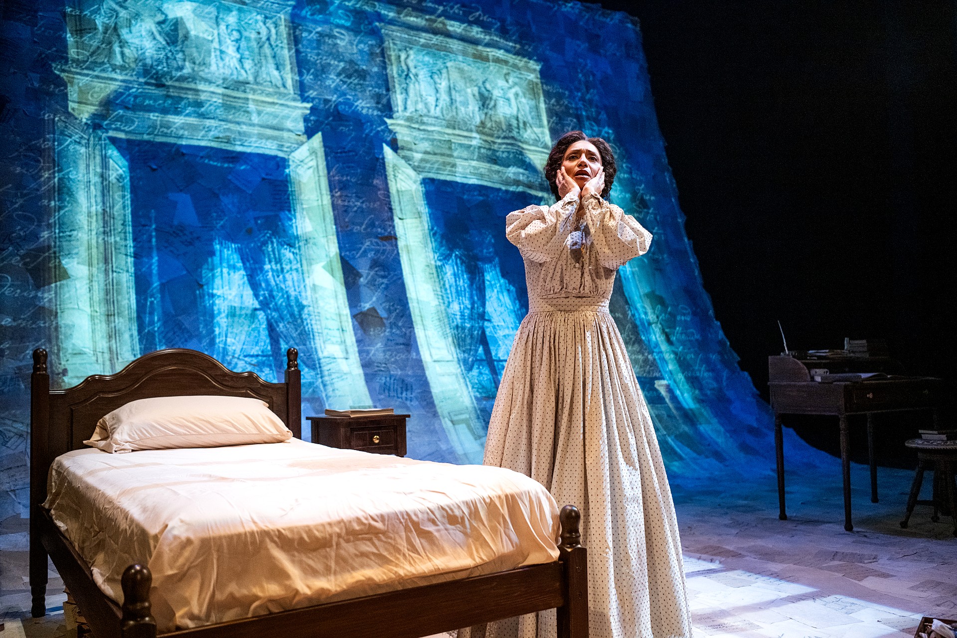 A wide stage shot showing a woman standing near a wooden bed with white linens and a simple frame. She is holding her face in her hands. The entire back wall is covered by a massive projection of blue-toned architectural doorways and handwritten letters.
